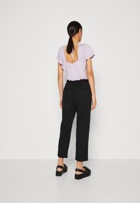 Blouse lilas avec un décolleté profond dans le dos, manches courtes et volantes. Associée à un pantalon noir ajusté et des sandales à plateforme noires. Texture lisse.