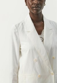 Hvid linned blazer med dobbeltbrystet front, klaplapper og to store knapper. Har en glat tekstur og skræddersyet pasform.