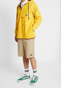 Yellow lightweight pullover jacket with a front pocket, black drawstring, beige shorts, and green sneakers with white accents.