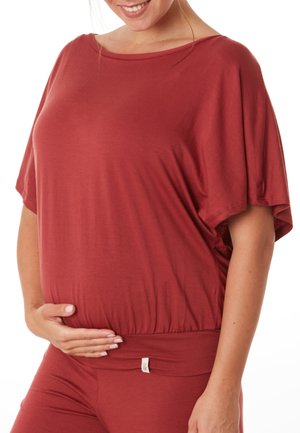 Ensemble de haut et de pantalon de maternité en couleur rouille. Le haut présente un décolleté large et des manches amples ; le bas a une taille élastique avec une petite étiquette.