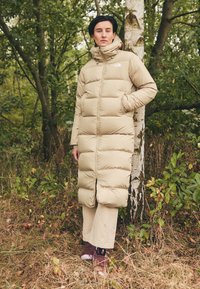 Parka beige, lunghezza ginocchio, design trapuntato, colletto alto, due tasche laterali. Abbinata a pantaloni chiari e sandali viola.