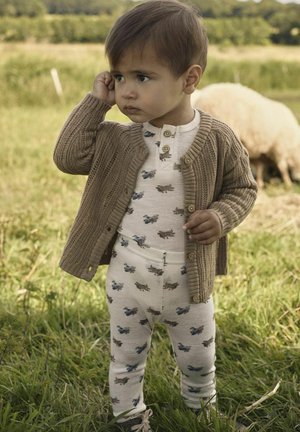 Brauner Strickcardigan über einem weißen Einteiler mit bunten Flugzeugmustern und passenden Hosen. Das Kind steht im Gras, die Hand ans Ohr.