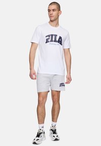 Camiseta blanca de manga corta con el logo de "FILA" en azul marino; pantalones cortos grises con un logo similar; zapatillas blancas con detalles y logo en negro; estilo casual.