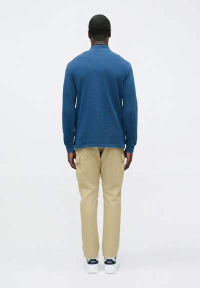 Modra majica z dolgimi rokavi in okroglim izrezom, z teksturirano površino, kombinirana s kaki hlačami s številnimi žepi in belimi supergami z mornarskimi poudarki.