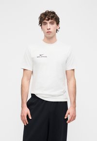 Balti medvilninė marškinėliai su apvaliu kaklu, priekyje juodas Nike swoosh logotipas ir užrašas „NIKE OFFICIAL“. Modelis vilki juodas kelnes.