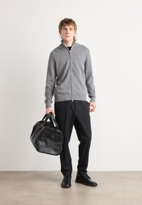 Gray zip-up sweater, black pants, and black shoes. Holding a black leather duffel bag with a rounded shape and prominent stitching detail.