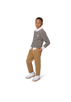 Un pull marron à motifs, une chemise blanche à boutons en dessous, un pantalon beige et des baskets blanches. Les détails notables incluent des poignets côtelés et un écusson sur le pull.