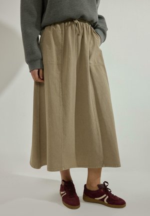 Person wearing a beige A-line skirt with an elastic waistband, gray sweatshirt, and maroon sneakers with white stripes, standing against a plain background.