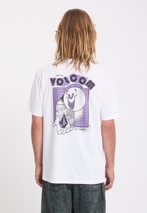 Personne aux cheveux longs portant un t-shirt blanc avec un graphisme violet d'un visage souriant et une main tenant une gemme, texte "Volcom Embrace The Strange."
