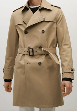 Mann trägt beige zweireihigen Trenchcoat mit schwarzen Knöpfen und Gürtel über schwarzem Pullover, dargestellt von Hals bis zur Mitte des Oberschenkels.