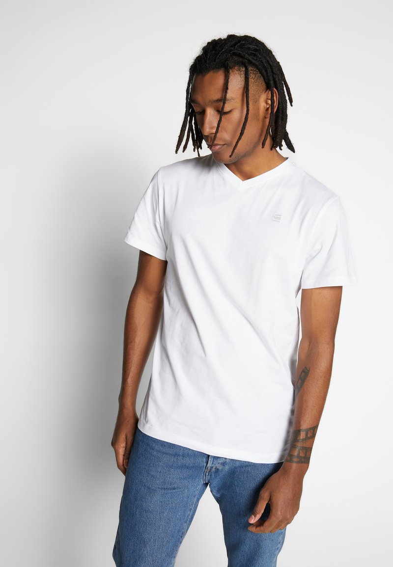 Weißes V-Ausschnitt-T-Shirt aus Baumwolle, mit kurzen Ärmeln und einem dezenten Logo auf der Brust; getragen mit blauen Jeans.