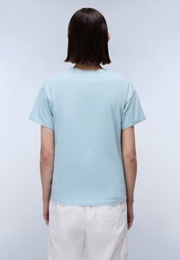 Lichtblauw katoenen T-shirt met korte mouwen, een relaxte pasvorm en een ronde hals, weergegeven vanaf de achterkant tegen een rustige achtergrond.