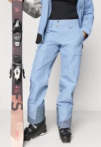 Ανοικτού μπλε σκι παντελόνι με λεία επιφάνεια, διαθέτοντας βαθιές τσέπες και αντιθέτον υφάσματος μπλε στο τελείωμα, συνδυασμένο με μαύρες μπότες σκι και σκι.