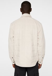 Ljusbeige ribbad jacka med en kviltad design, knäppta ärmslut och rundad nederkant. Texturerade ränder löper vertikalt över tyget.