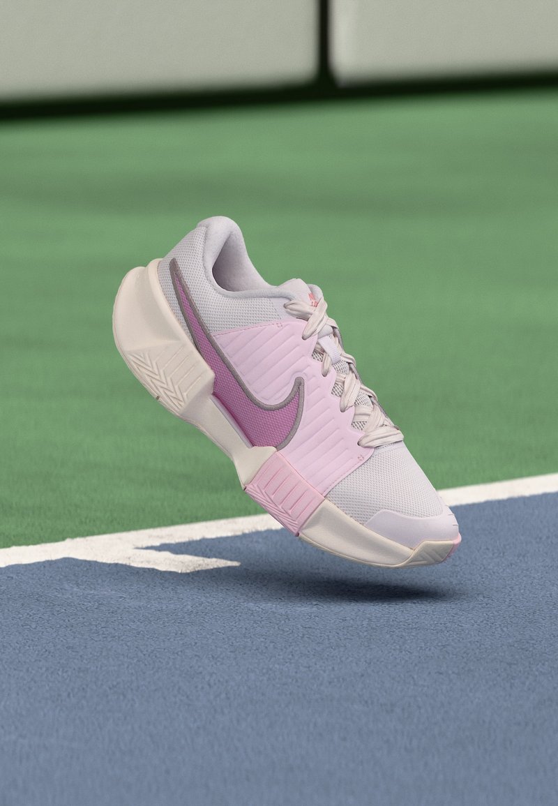 Ανοιχτό ροζ και λευκό αθλητικό παπούτσι Nike με μωβ λογότυπο να αιωρείται πάνω από το μπλε και πράσινο γήπεδο τένις.