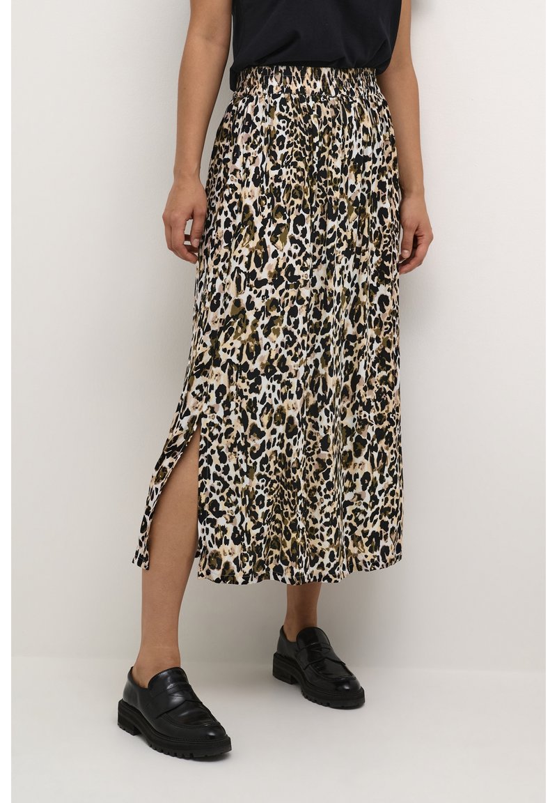 Femme portant une jupe midi imprimé léopard avec une fente latérale, un haut noir et des mocassins en cuir noir, debout devant un fond uni.