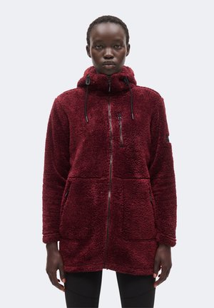 Personne portant une longue veste polaire bordeaux zippée avec capuche et cordons, debout devant un fond clair uni.