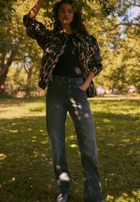 Chaqueta de leopardo con un corte holgado, debajo una camiseta negra y jeans de tiro alto en azul oscuro. Escenario exterior con vegetación de fondo.