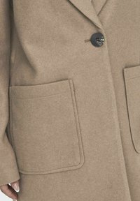 Abrigo de mezcla de lana beige con solapa de muesca, cierre de un solo botón negro y dos bolsillos frontales. Textura suave con detalles de costura limpios.