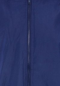 Tamsiai mėlyna striukė su pilnu ilgumo užtrauktuku per vidurį. Medžiaga atrodo lygi su šiek tiek blizgesio. Nėra matomų raštų ar dekoracijų.