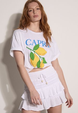 Rothaarige Frau trägt ein weißes, kurzes T-Shirt mit „CAPRI“- und Zitronen-Aufdruck, kombiniert mit einem weißen Minirock mit Rüschen und Kordelzug in der Taille.