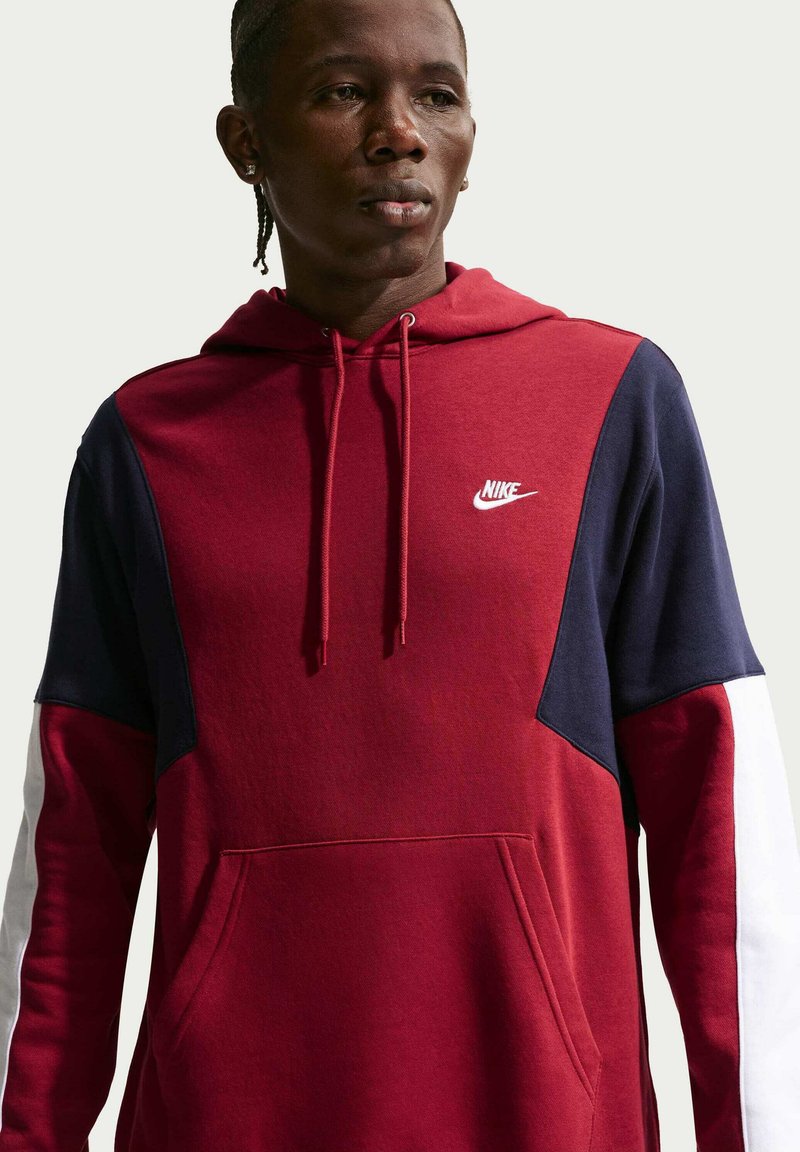Uomo con felpa con cappuccio Nike bordeaux, blu navy e bianca con tasca frontale, in piedi davanti a uno sfondo chiaro e semplice, che guarda di lato.