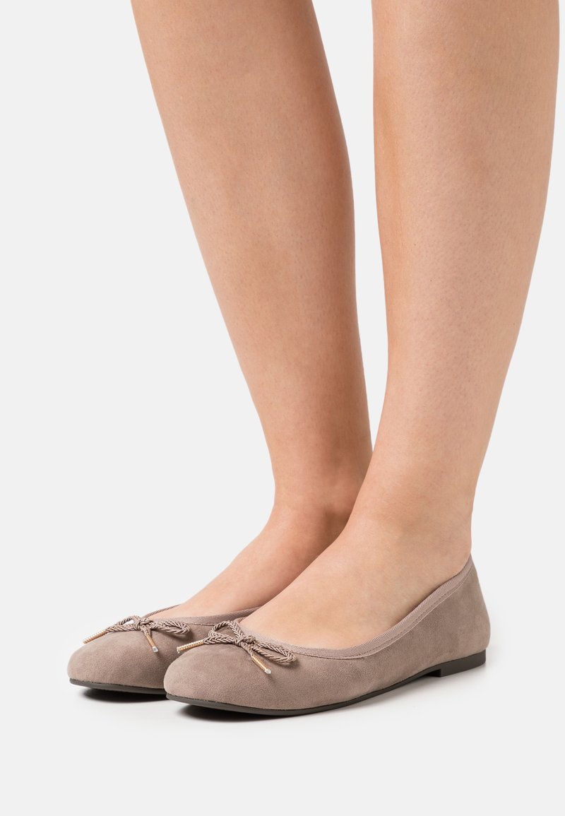 Ballerines en daim beige avec nœud décoratif et extrémités dorées portées pieds nus sur un fond clair uni.