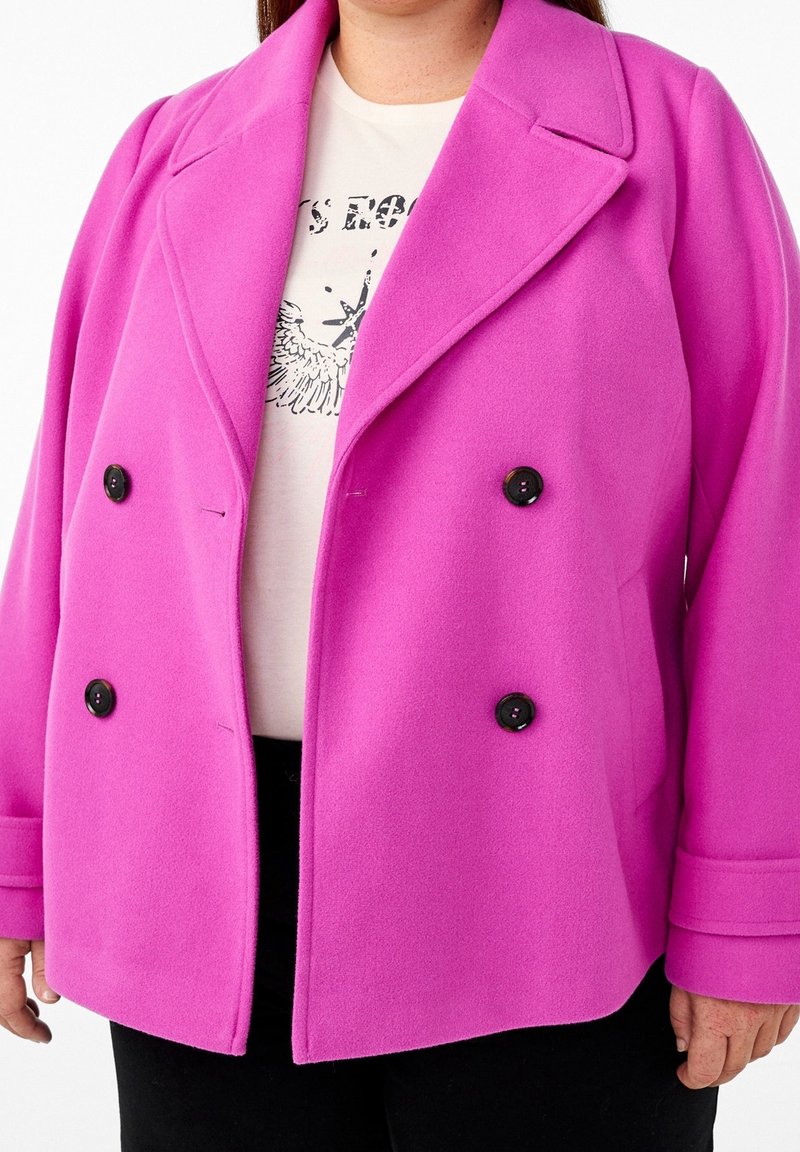 Personne portant un manteau croisé rose vif par-dessus un t-shirt blanc graphique et un pantalon noir, debout devant un fond uni.
