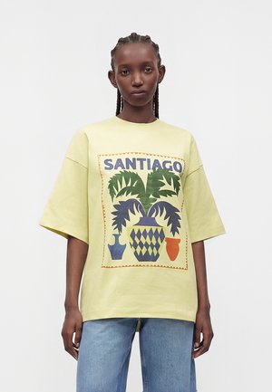 Ung person med flettet hår har en stor bleg gul T-shirt med teksten "SANTIAGO" og farverig grafik af et blad og en vase, kombineret med blå jeans.