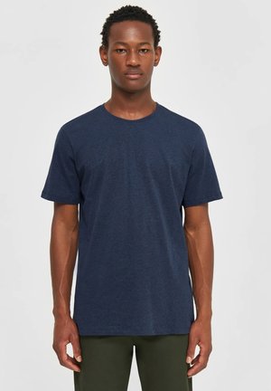 Giovane uomo in piedi di fronte, indossa una maglietta blu scuro a maniche corte tinta unita e pantaloni verde oliva su sfondo neutro.