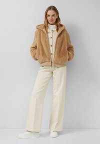 Beige nep-bont met capuchon, met een ritssluiting aan de voorkant, geribbelde manchetten en onderrand. G gedragen over een crèmekleurige gebreide top en gecombineerd met crèmekleurige wijde broeken en witte sneakers.
