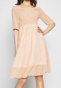 Žena v světle broskvovém, šaty po kolena s krátkými rukávy a nabranou, smockovanou hrudí, s jednou rukou položenou na boku.