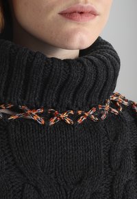 Tjock svart stickad tröja med hög ribbad krage, med färgglada flätade detaljer längs halsringningen. Texturerad garninformation.