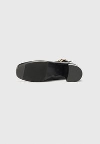 Semelle de chaussure en cuir noir présentant une surface lisse, des rainures horizontales pour l'adhérence, et un talon arrondi. Détail décoratif en métal doré visible.