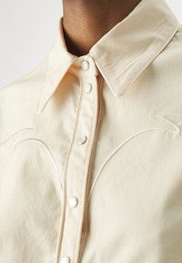 Lichtbeige shirt met een puntkraag, voorzien van knoopsluitingen en subtiele witte geborduurde accenten op de borst. Gladde textuur.