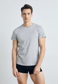 T-shirt gris à manches courtes en mélange de coton, avec un col rond et une coupe décontractée. Le modèle se tient devant un fond uni.