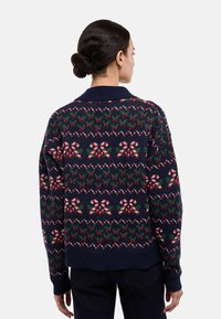 Cardigan en maille marine avec des motifs festifs rouges et verts, comprenant des cannes de bonbon et des feuilles, avec un col et des poignets côtelés.