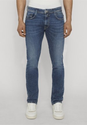 Man draagt slim-fit blauwe spijkerbroek met lichte vervaging en witte sneakers, staande tegen een eenvoudige lichte achtergrond.