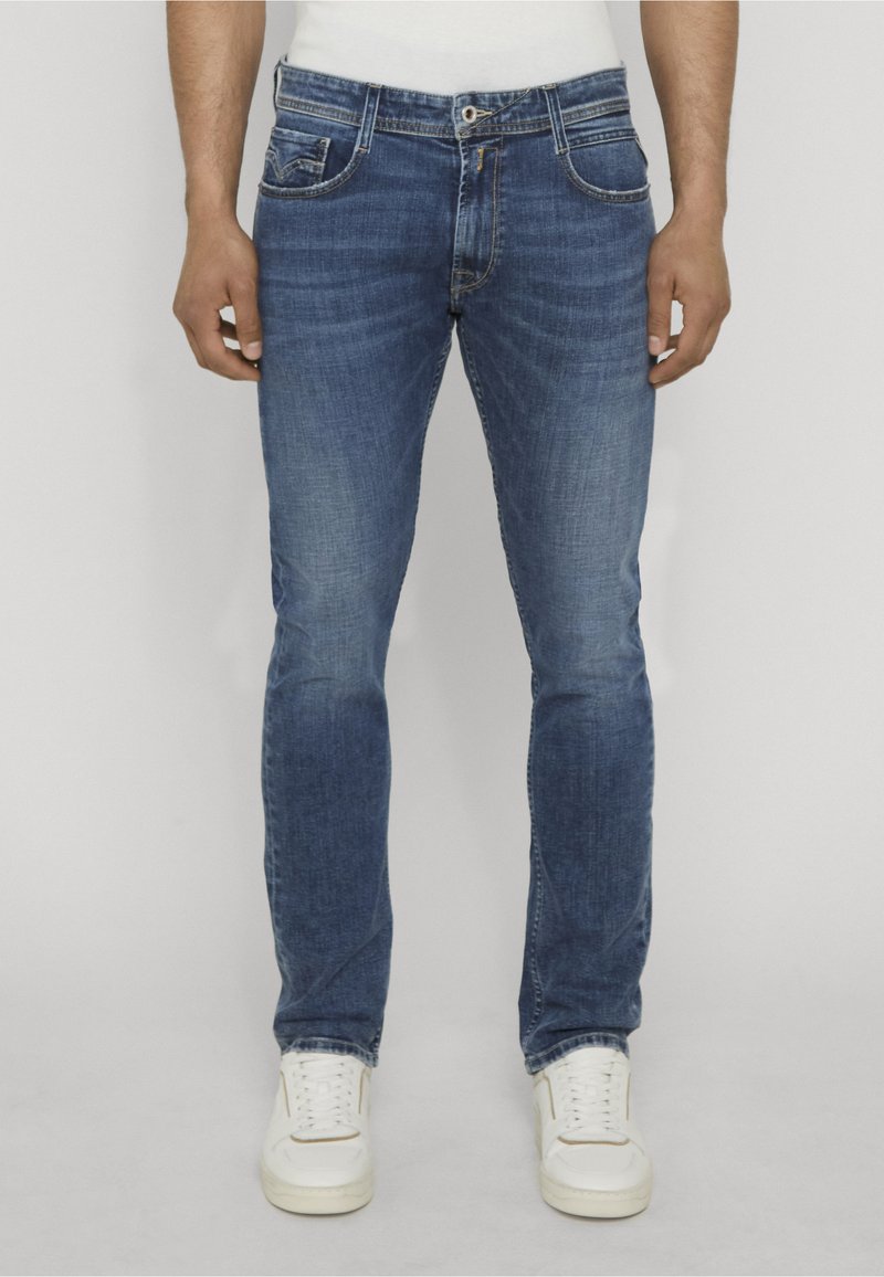 Homme portant un jean bleu slim légèrement délavé et des baskets blanches, debout contre un fond clair uni.