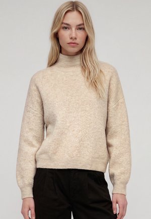 Femme blonde portant un pull à col roulé beige et un pantalon noir, debout devant un fond clair uni.
