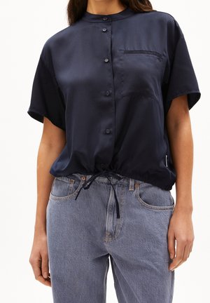 Kobieta ubrana w granatową bluzkę na guziki z krótkim rękawem, z kieszenią na piersi i ściągaczem na dole, w połączeniu z jasnoniebieskimi dżinsami.