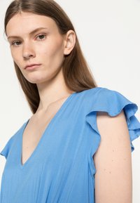 Vestido azul com decote em V e mangas de flutter, feito de um tecido suave. A textura parece lisa, com um caimento leve e mínimas tranças.