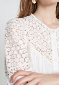 Biała bluzka z misternymi koronkowymi zdobieniami na rękawach, zapinana na guziki, o plisowanym fasonie. Tkanina jest lekka i delikatna.