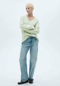 Hellgrüner, locker sitzender Pullover mit V-Ausschnitt, kombiniert mit hellblauen, weit geschnittenen Jeans und schwarzen Schuhen, vor einem neutralen Hintergrund.