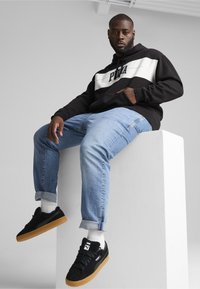 Sudadera negra con franjas blancas y logo, vaqueros azul claro con dobladillos, zapatillas negras con suela de goma y calcetines blancos, sentado en un pedestal.