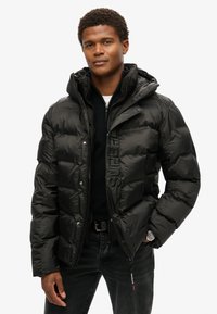 Sort pufferjakke med glansfinish, lynlås foran, hætte og broderet branding på brystet. Båret over en mørk sweater og jeans.