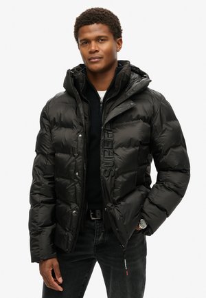 Chaqueta tipo puffer negra con acabado brillante, cierre frontal con cremallera, capucha y logotipo bordado en el pecho. Lleva puesta sobre un suéter oscuro y jeans.
