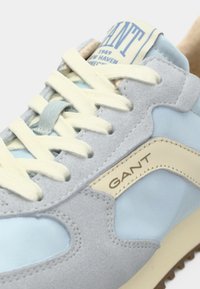 Ανοιχτό μπλε και γκρι αθλητικό παπούτσι με κρεμ κορδόνια και μπεζ ετικέτα λογότυπου που γράφει "GANT", παρουσιάζοντας λεπτομερείς ραφές και υφασμάτινες υφές.