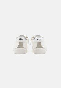 Baskets en cuir blanc avec des accents en daim gris sur le talon, présentant un logo à l'arrière, une semelle en caoutchouc plate et un design minimaliste.