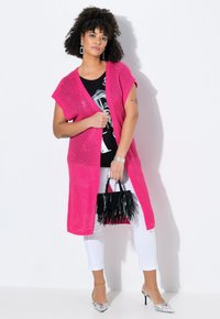 Rosa, offener Strickcardigan mit kurzen Ärmeln, getragen über einem schwarzen Grafik-T-Shirt, kombiniert mit weißen Hosen und einer schwarzen, gefransten Handtasche.